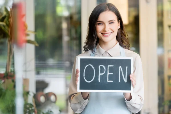 Happy florist holding board with open lettering near flower shop while looking at camera — Stock Photo