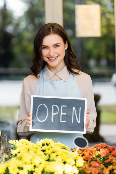 Fiorista felice sorridente alla macchina fotografica mentre tiene bordo con scritte aperte vicino crisantemi su primo piano sfocato — Foto stock