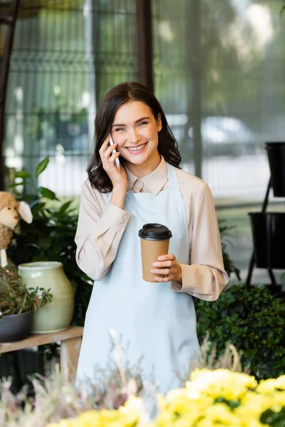 Fiorista allegro parlando su smartphone e tenendo il caffè per andare vicino fiori sfocati in primo piano — Foto stock