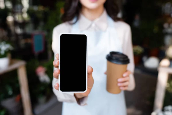 Vista recortada de floristería sosteniendo teléfono inteligente con pantalla en blanco y café para ir durante el freno sobre fondo borroso - foto de stock