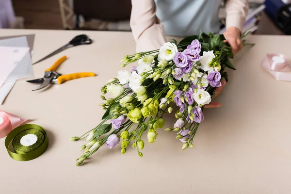Partial view of florist holding eustoma flowers near decorative tape, secateurs and scissors on blurred background — Stock Photo