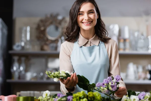 Fröhlicher Blumenhändler mit Eustoma-Blumen, während er auf verschwommenem Vordergrund in die Kamera lächelt — Stockfoto