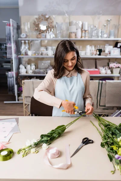 Smiling florist cutting steam while making eustoma flowers bouquet near rack with vases on blurred background — Stock Photo