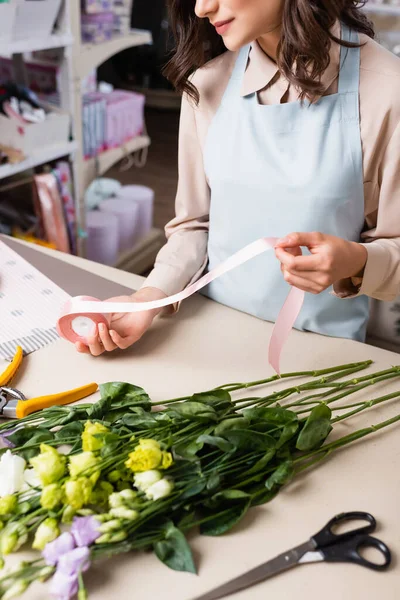 Vista parcial do florista segurando fita decorativa ao fazer buquê de flores eustoma na loja de flores em primeiro plano borrado — Fotografia de Stock