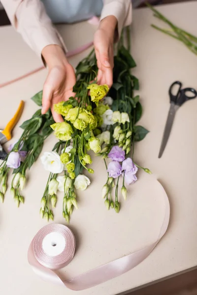 Vue de dessus du fleuriste tenant des fleurs d'eustome près des ciseaux et du ruban décoratif tout en faisant un bouquet dans la boutique de fleurs — Photo de stock