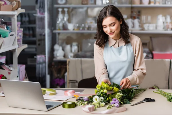 Young florist making bouquet with eustoma flowers near laptop and racks on blurred background — Stock Photo