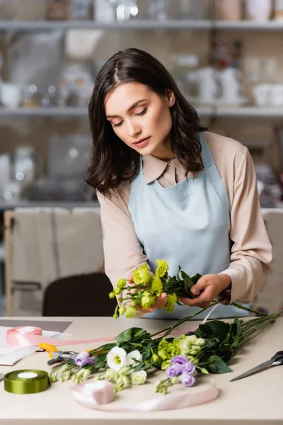 Young florist arranging bouquet with eustoma flowers near decorative ribbon on blurred foreground — Stock Photo