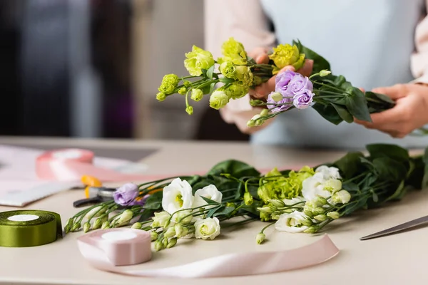 Vista cortada de florista segurando flores eustoma perto de fita decorativa na mesa sobre fundo borrado — Fotografia de Stock