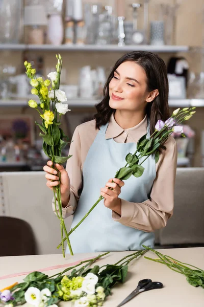 Florista feliz haciendo ramo con flores eustoma cerca de rack con jarrones sobre fondo borroso - foto de stock