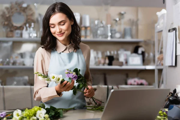 Fiorista sorridente che tiene fiori di eustoma vicino al computer portatile in primo piano sfocato — Foto stock