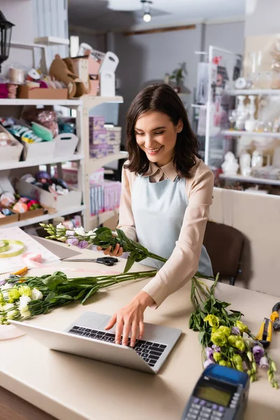 Fröhliche Floristin mit Laptop, während sie Eustoma-Blumen in der Nähe von Regalen auf verschwommenem Hintergrund hält — Stockfoto