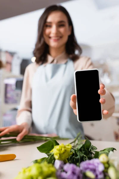 Vista de cerca del teléfono inteligente en la mano de florista sonriente de pie cerca del escritorio con flores de eustoma sobre un fondo borroso - foto de stock