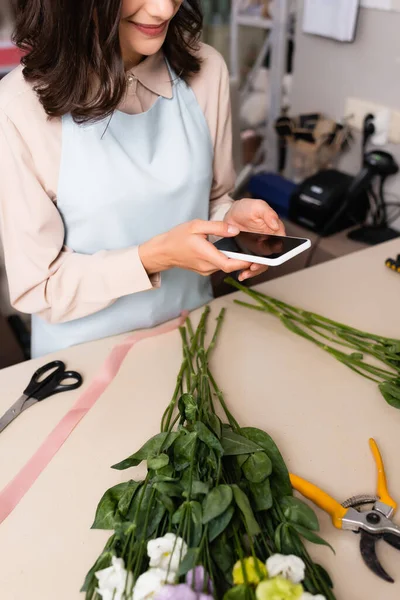 Cropped view of florist texting on smartphone, while standing near desk with eustoma flowers on blurred background — Stock Photo