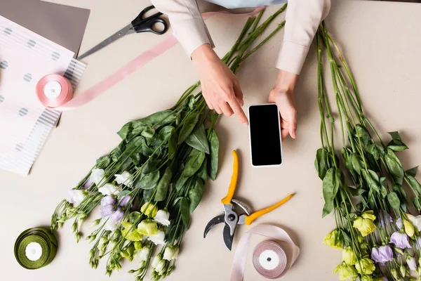 Ausgeschnittene Ansicht eines Blumenhändlers, der mit dem Finger auf sein Smartphone zeigt, auf dem Schreibtisch mit dekorativen Bändern, Werkzeugen und Eustoma-Blumen — Stockfoto