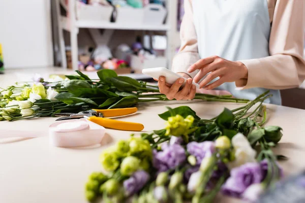Vista recortada de la floristería con teléfono inteligente cerca del escritorio con flores de eustoma borrosas en primer plano - foto de stock