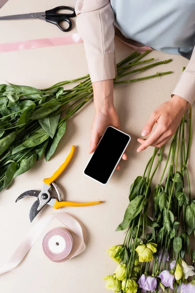 Vue recadrée du fleuriste tenant smartphone avec écran vierge près des rubans décoratifs, des outils et des fleurs d'eustomie om bureau — Photo de stock