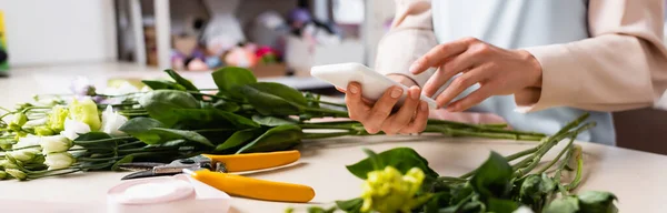 Cropped view of florist using cellphone near decorative ribbon, secateurs and eustoma flowers on desk on blurred background, banner — Stock Photo