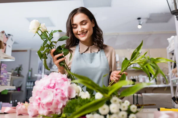 Glückliche Floristin komponiert Blumenstrauß mit Pflanze und Rosen mit verschwommenen Blumen im Vordergrund — Stockfoto