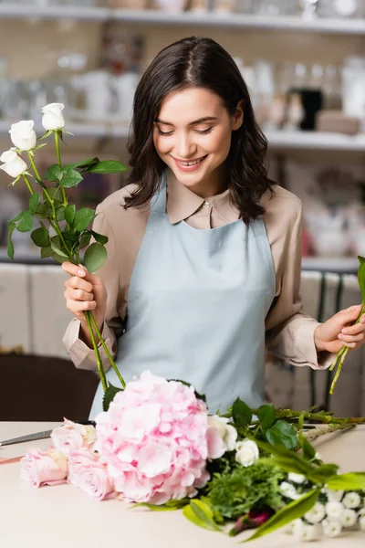 Joyeux fleuriste féminin avec des roses regardant des fleurs sur le bureau, tout en composant un bouquet sur fond flou — Photo de stock