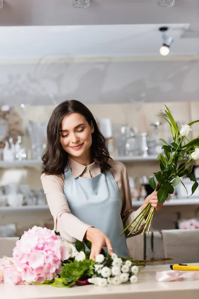 Sorridente fiorista bruna con fiori che prendono crisantemi dalla scrivania con rastrelliere sfocate sullo sfondo — Foto stock