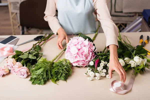 Cropped view of florist taking decorative ribbon near flowers on desk with tools on blurred background — Stock Photo