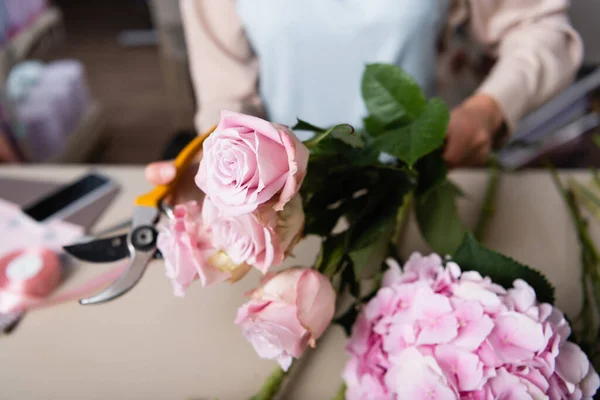 Close up view of roses and hydrangea in hand of florist holding secateurs with blurred desk on background — Stock Photo