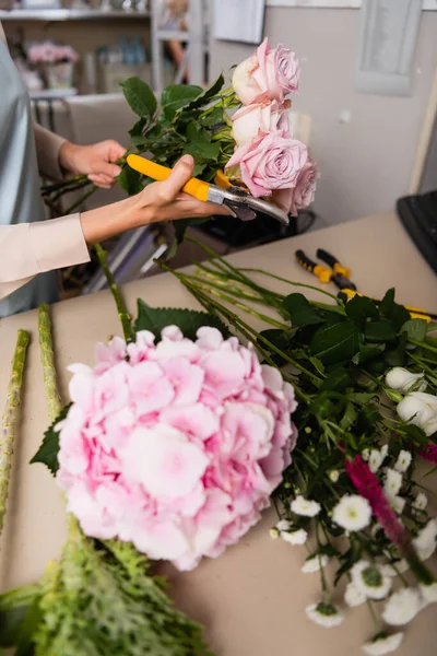 Cropped view of florist with secateurs and roses standing near desk with blurred flowers on foreground — Stock Photo