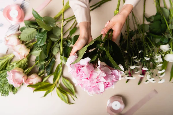 Vista ritagliata del fiorista che tiene ortensia in fiore, mentre compone mazzi di fiori vicino a fiori e nastri decorativi sulla scrivania — Foto stock