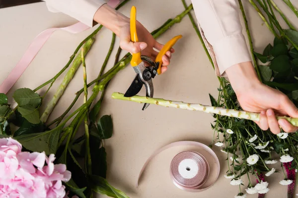 Vista ritagliata di fiorista con forbici fusto di taglio di pianta vicino a fiori e nastri decorativi sulla scrivania — Foto stock