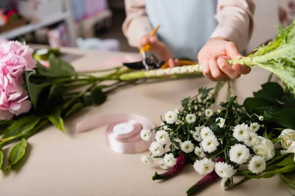 Vue recadrée du fleuriste avec sécateurs coupant la tige de la plante près des fleurs et le ruban décoratif sur le bureau sur fond flou — Photo de stock