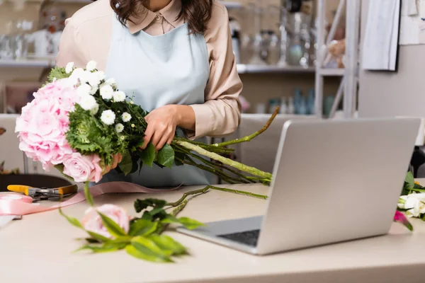 Vista recortada de floristería que arregla ramo con rosas, crisantemos y hortensias cerca de la computadora portátil en el escritorio sobre fondo borroso - foto de stock