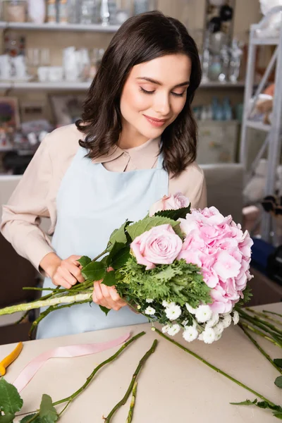 Sonriente florista morena mirando ramo cerca de escritorio con tallos en bastidores borrosos en el fondo - foto de stock