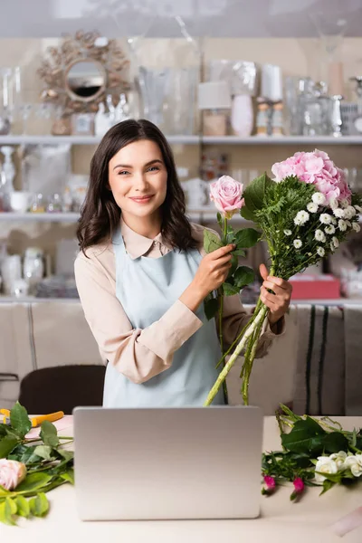 Florista femenina sonriente mirando a la cámara mientras compone ramo cerca de la computadora portátil en el escritorio con bastidores borrosos en el fondo - foto de stock