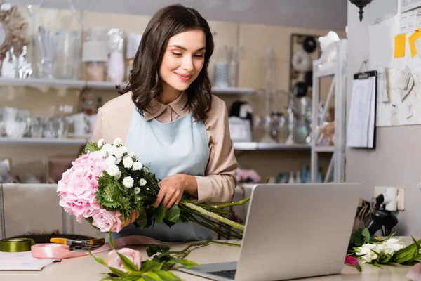 Fleuriste souriante regardant ordinateur portable, tout en arrangeant un bouquet avec des rayonnages flous sur le fond — Photo de stock