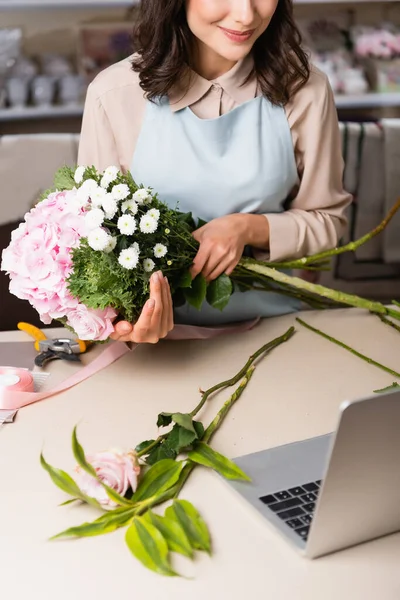 Vue recadrée du bouquet de fleuriste tenant avec hortensia, rose et chrysanthèmes près de l'ordinateur portable sur le bureau sur fond flou — Photo de stock