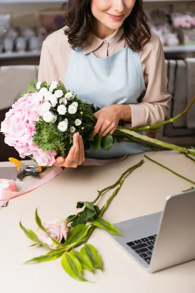 Cropped view of florist holding bouquet with hydrangea, rose and chrysanthemums near laptop on desk on blurred background — Stock Photo