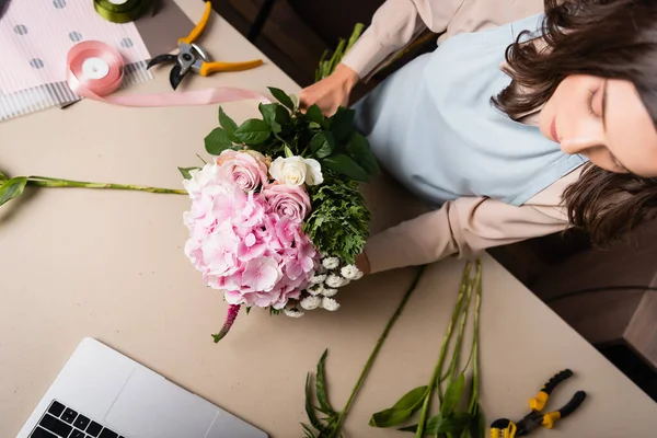 Overhead view of brunette florist arranging bouquet on desk with tools, wrapping papers and decorative ribbons — Stock Photo