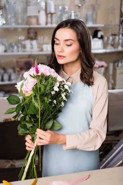 Fleuriste féminine focalisée arrangeant bouquet près du bureau avec des rayonnages flous de vases sur fond — Photo de stock