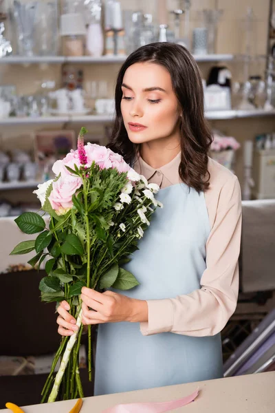 Konzentrierte Floristin arrangiert Blumenstrauß in der Nähe von Schreibtisch mit verschwommenen Regalen von Vasen auf dem Hintergrund — Stockfoto