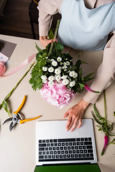 Cropped view of female florist using laptop, while holding bouquet on desk with secateurs and decorative ribbon — Stock Photo