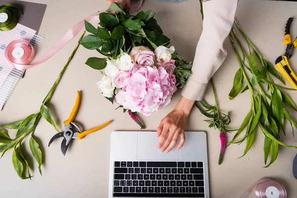 Vue recadrée du fleuriste féminin à l'aide d'un ordinateur portable, tout en tenant un bouquet sur le bureau avec des outils, des plantes et des rubans décoratifs — Photo de stock
