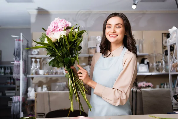 Happy female florist looking at camera, while arranging bouquet with blurred racks of vases on background — Stock Photo