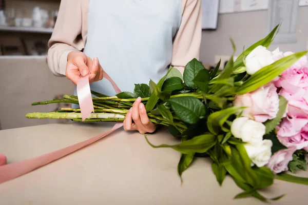Cropped view of female florist with decorative ribbon tying stalks of fresh bouquet in flower shop on blurred background — Stock Photo