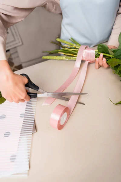 Vista recortada de floristería con tijeras cortando cinta decorativa cerca de tallos atados de ramo en escritorio - foto de stock