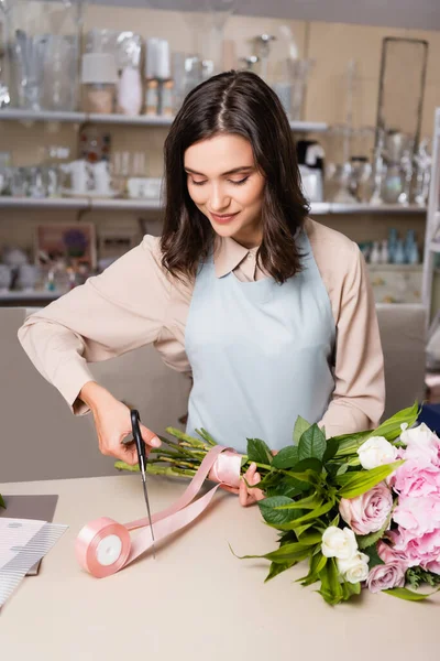 Fleuriste féminine positive avec ciseaux coupant ruban décoratif, tout en tenant bouquet attaché avec des rayonnages flous sur le fond — Photo de stock