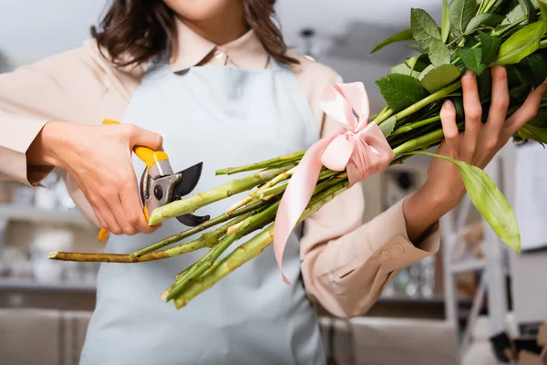 Cropped view of female florist with secateurs cutting stalks of tied bouquet on blurred background — Stock Photo