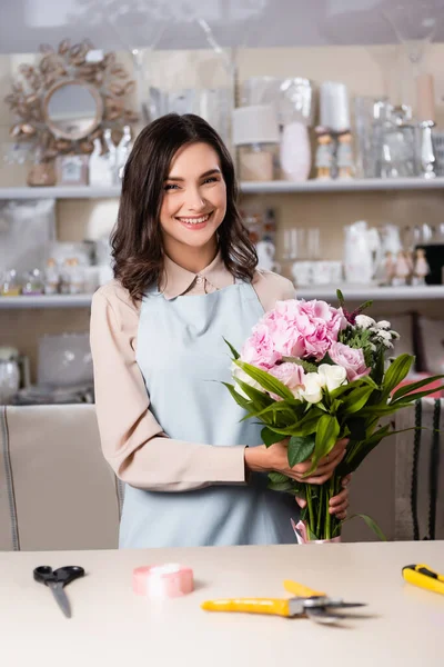 Cheerful florist holding bouquet near desk with tools and decorative ribbon on blurred background — Stock Photo