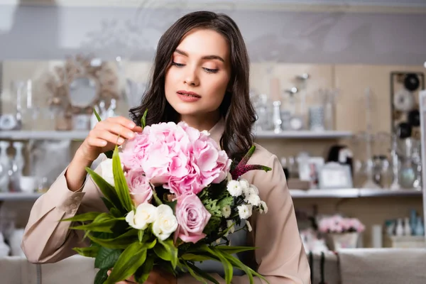 Brunette fleuriste féminine touchant hortensia floraison, tout en regardant le bouquet avec des rayonnages flous de vases sur fond — Photo de stock