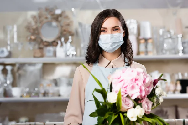 Brunette fleuriste en masque médical avec bouquet dans la boutique de fleurs avec des rayonnages flous sur le fond — Photo de stock