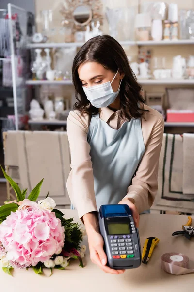 Fleuriste féminine dans le masque médical tenant terminal, tout en regardant le bouquet sur le bureau sur fond flou — Photo de stock